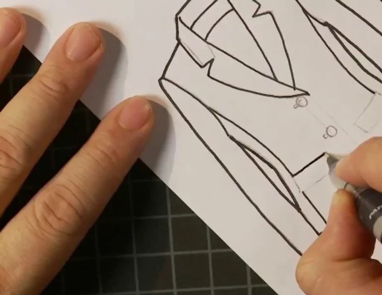 A person draws a black outline of a suit jacket on white paper, using a pen. The drawing is on a gridded black cutting mat.