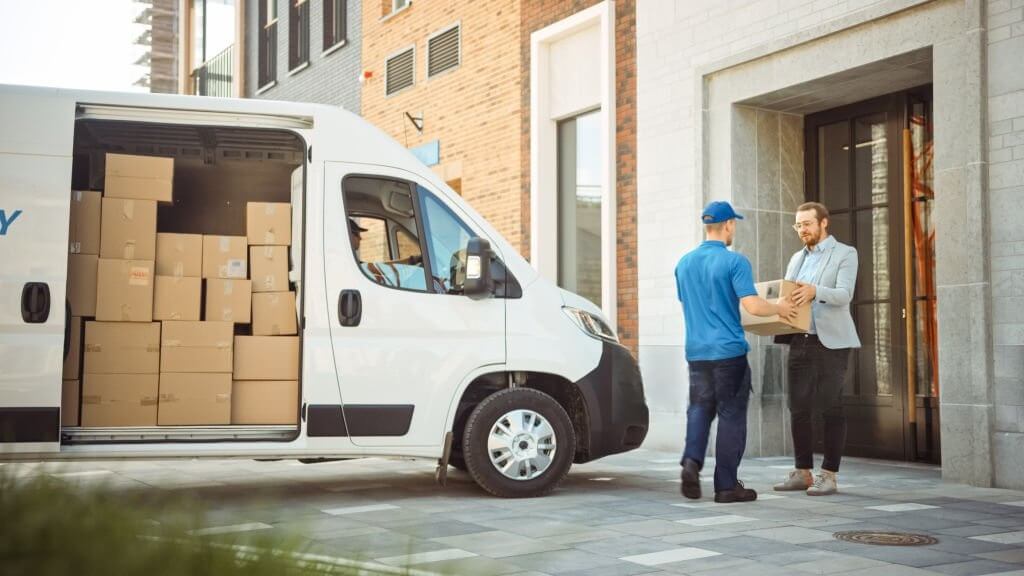 A delivery worker hands a package to a man outside a building, with a white van full of cardboard boxes parked nearby.